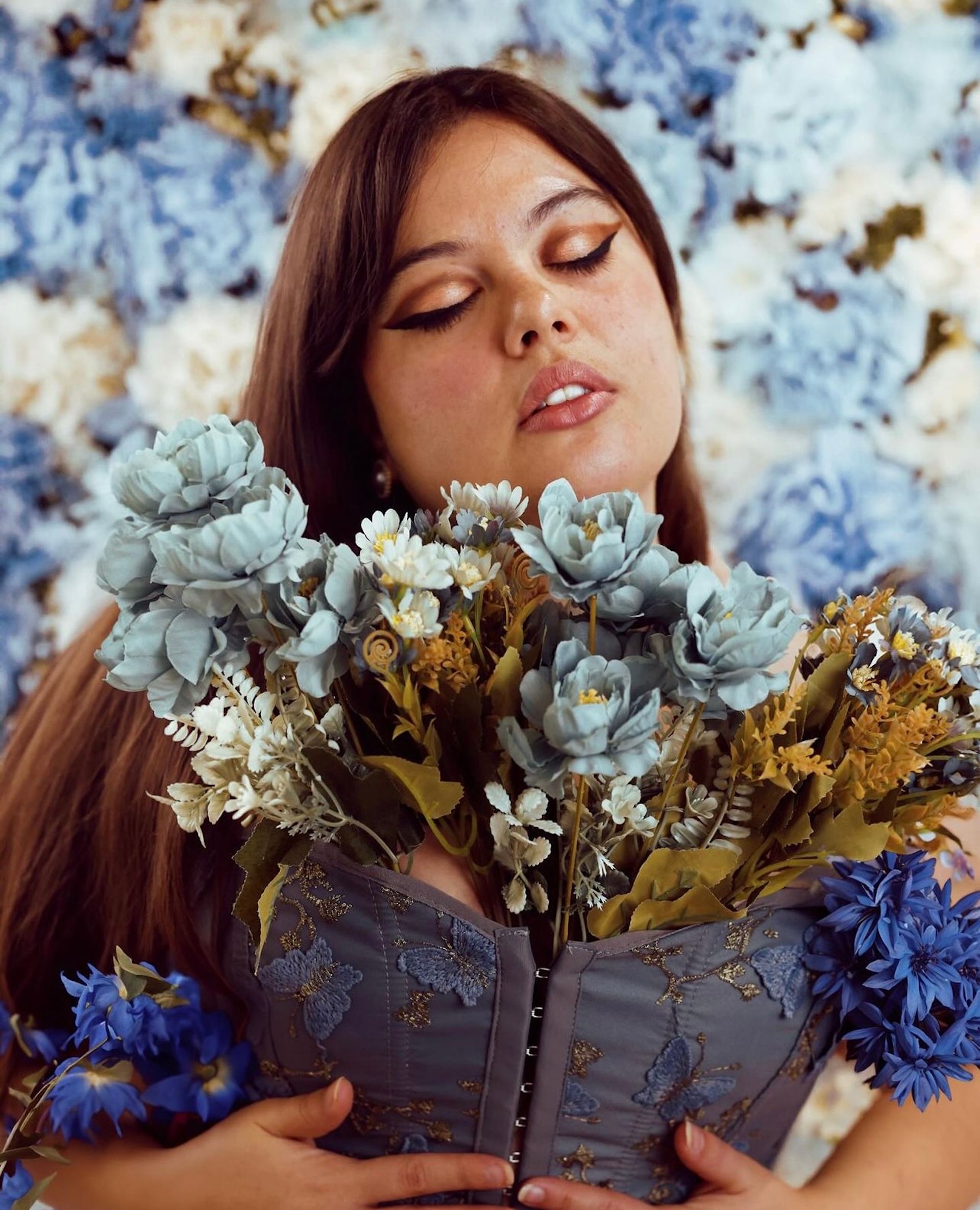 Adriana May, con los ojos cerrados, sostiene un ramo de flores azules y blancas frente a una pared de hortensias azules